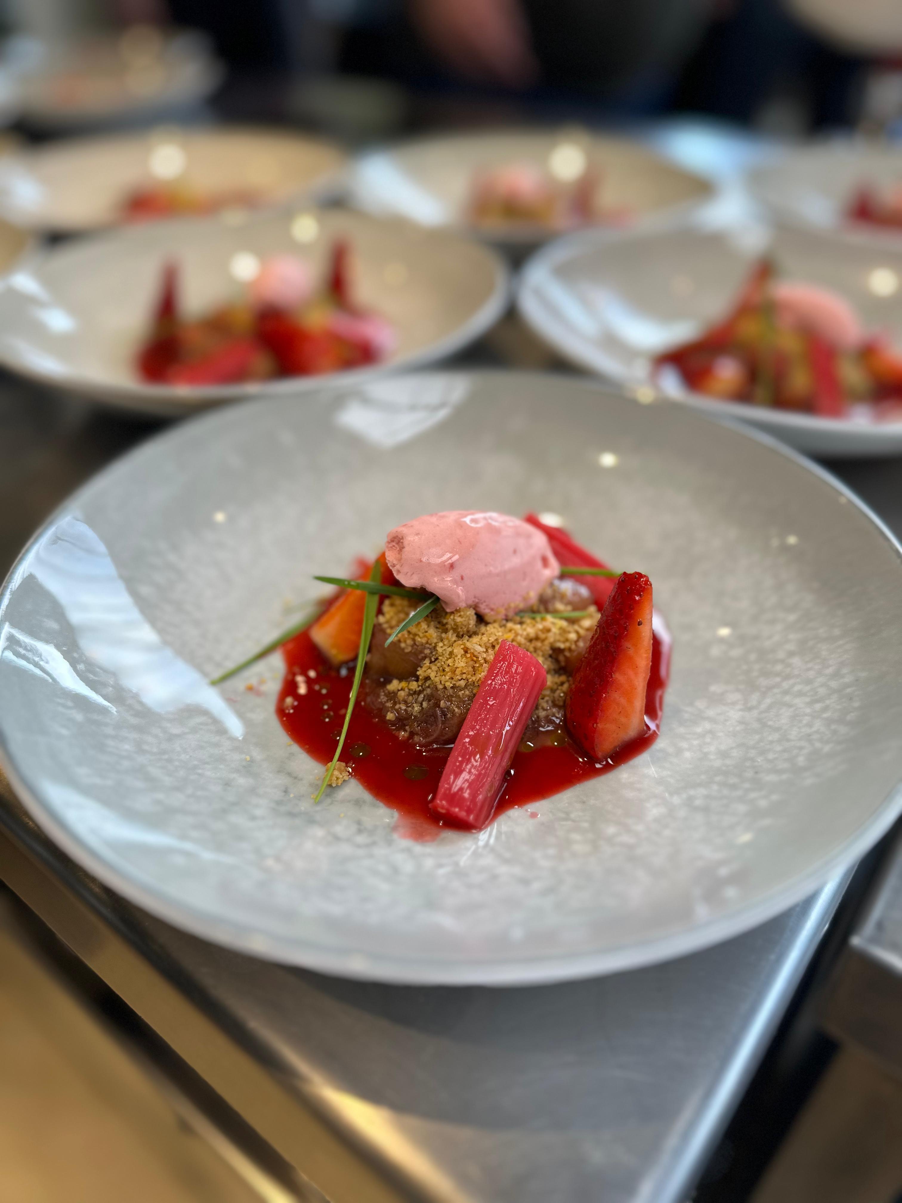 Teller mit Frühlingsdessert aus Rhabarber, frischen Erdbeeren, Streuseln und rosa Sorbet – angerichtet auf einem hellgrauen Teller, im Hintergrund weitere identische Desserts