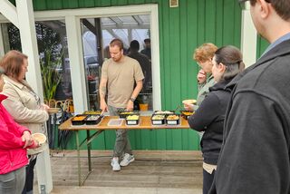 Person bereitet an einer mobilen Kochstation verschiedene Gerichte mit regionalen Zutaten vor, umgeben von interessierten Teilnehmern auf einer überdachten Terrasse vor einem grünen Gebäude