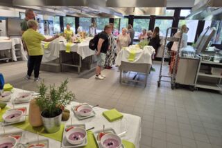 Besucherinnen und Besucher der Gesundheitswoche im bfw Hamm versammeln sich in der Showküche, in der verschiedene Kochstationen vorbereitet sind – im Vordergrund dekorativ angerichtete Speisen und frische Kräuter.