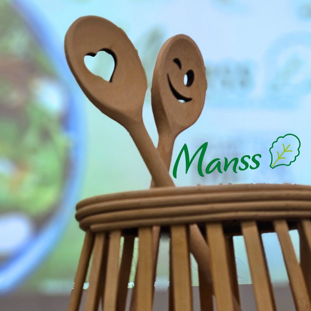 Zwei Holzlöffel mit Herz- und Smiley-Ausschnitt in einem Holzgefäß vor unscharfem Hintergrund mit Manss-Logo