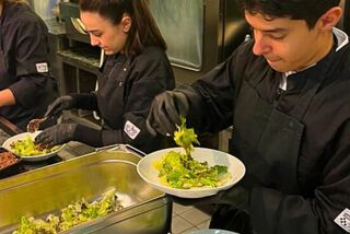 Zwei Mitarbeitende in schwarzer Berufskleidung richten frischen Salat in Bowls an, im Hintergrund eine Großküche.