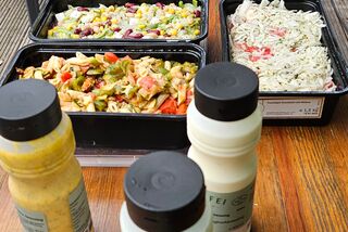 Auf einem Holztisch stehen mehrere Behälter mit verschiedenen Salaten und Beilagen wie Nudelsalat, Krautsalat, Linsensalat und Kichererbsen – im Vordergrund stehen mehrere Flaschen mit Dressings, im Hintergrund ist eine grüne Außenwand zu sehen