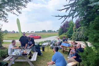 Teilnehmende der Manss Foodservice Produktschulung sitzen im Schatten unter Bäumen, während eine Produktvorstellung im Freien auf dem Hof stattfindet – im Hintergrund Felder, ein rotes Auto und ländliche Atmosphäre