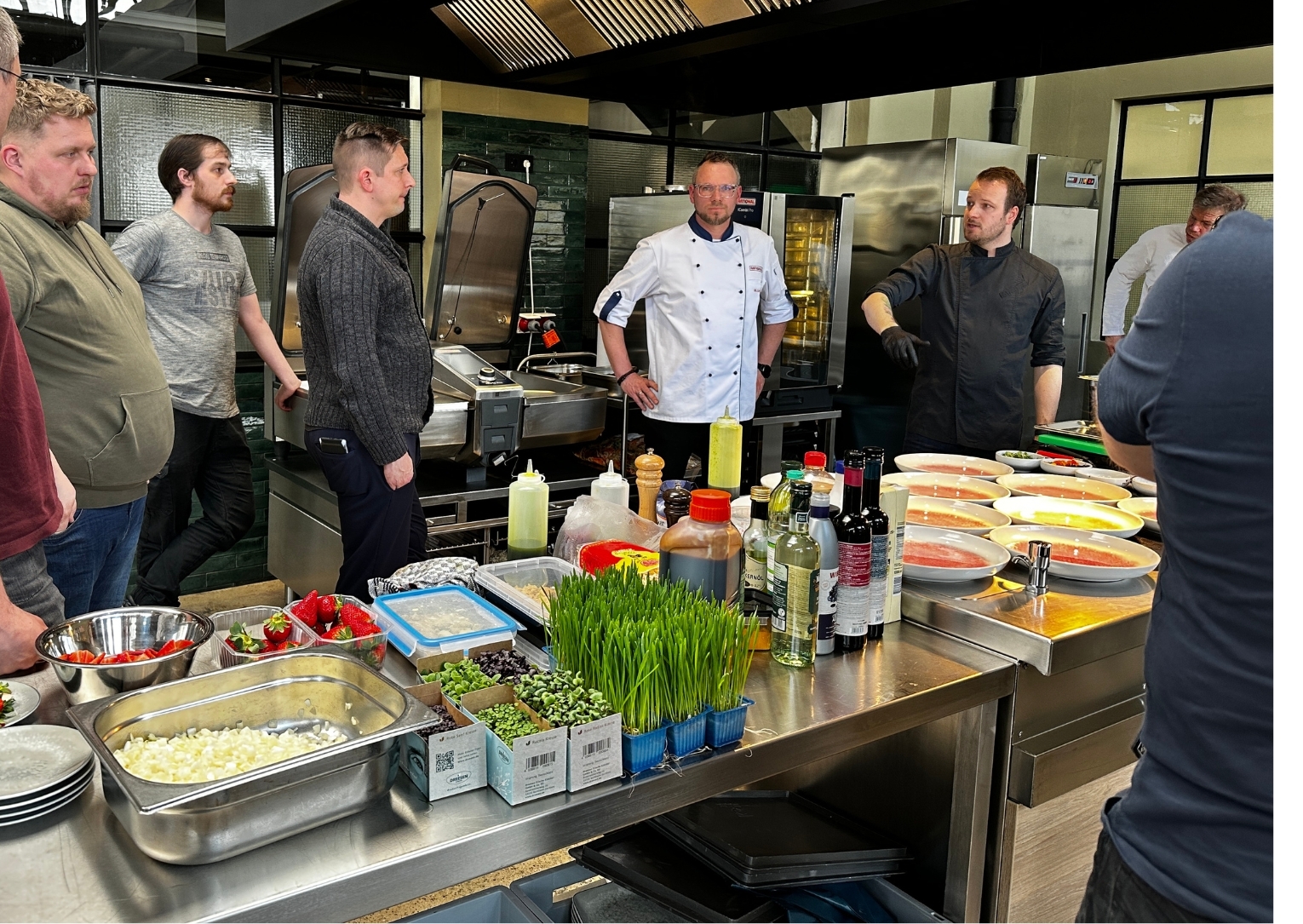 Zwei Köche im Gespräch mit Workshop-Teilnehmenden in der Showküche von Manss Foodservice – frische Zutaten, Teller und Küchenutensilien auf der Arbeitsfläche im Vordergrund, Edelstahlgeräte und Ofen im Hintergrund