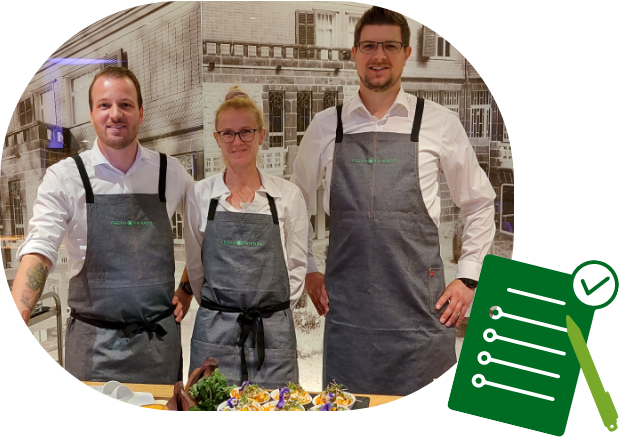 Drei Mitarbeitende im einheitlichen Outfit mit Schürzen vor einem dekorierten Stand – bereit für ihren Einsatz bei einer Veranstaltung mit frischen Produkten.