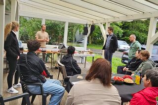 Teilnehmende eines Kochkurses sitzen unter einer überdachten Terrasse in einer Abschlussrunde, während ein Kursleiter spricht – im Hintergrund frisches Obst und Gemüse