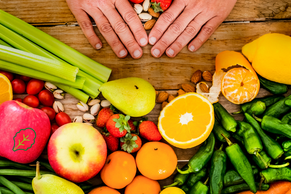 Bunte Auswahl an frischem Obst und Gemüse wie Äpfel, Orangen, Karotten, Sellerie, Paprika, Erdbeeren und Tomaten auf einem Holztisch. Zwei Hände formen ein Herz über einer Handvoll Beeren – Symbol für Liebe zu regionaler und nachhaltiger Ernährung.
