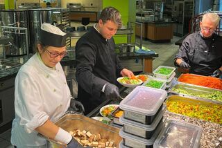 Drei Mitarbeitende bereiten in einer Großküche bunte Bowls mit frischen Zutaten vor; im Vordergrund mehrere Boxen mit geschnittenem Gemüse.