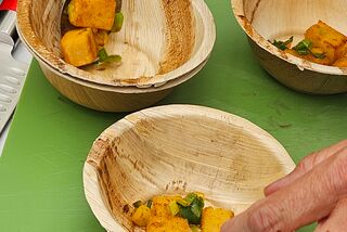 Hände richten in kleinen Palmblattschalen ein Gericht mit gebratenem Kürbis und Kräutern an, im Hintergrund liegen Kochutensilien auf einer grünen Unterlage