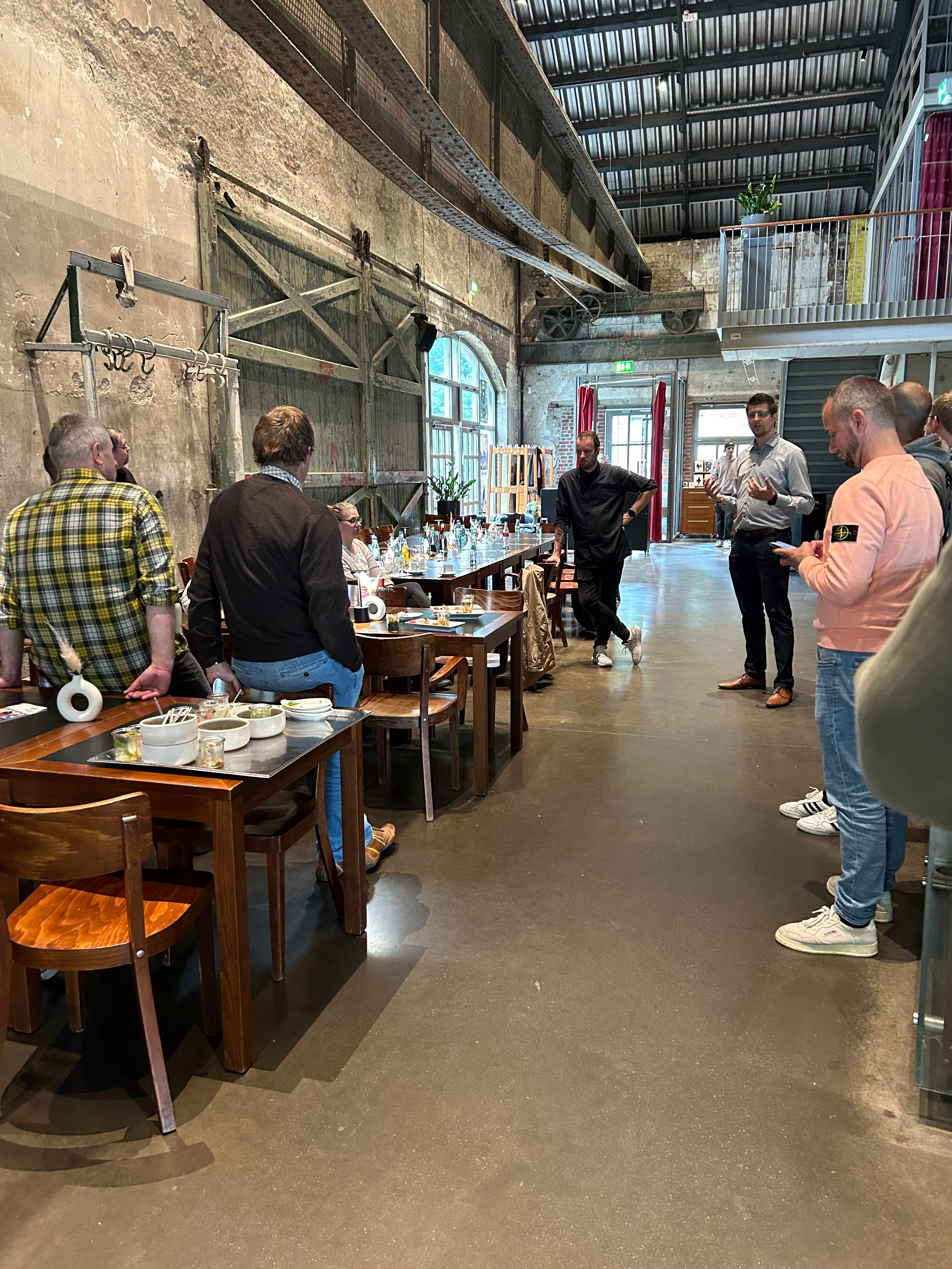 Teilnehmende stehen und sitzen in rustikaler Halle, Einführung durch Workshop-Leitung