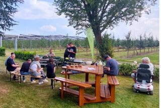 Mitarbeiter von Manss Foodservice sitzen bei einer Produktschulung im Grünen auf Bänken und Picknicktischen, mit Blick auf Felder und Obstplantagen.