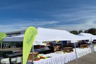 Weiß eingedeckte Marktstände mit frischem Obst und Gemüse beim Sommerfest von Blumen Risse und Overkamp – im Vordergrund ein grüner Manss-Banner