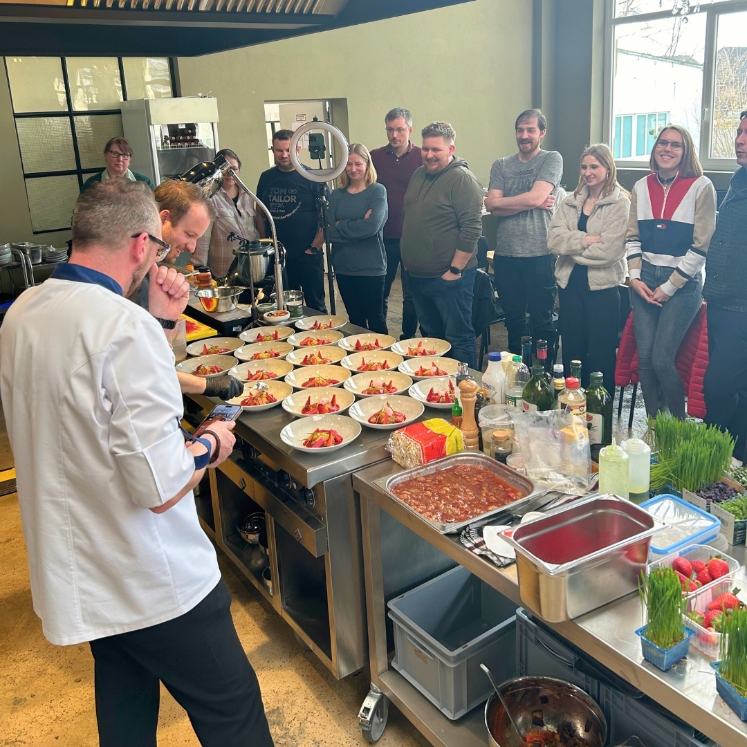 Köche bereiten beim Manss-Workshop in der Showküche das Rhabarber-Dessert zu – mehrere angerichtete Teller auf der Arbeitsfläche, umgeben von Workshop-Teilnehmenden, frische Zutaten und Geräte im Vordergrund