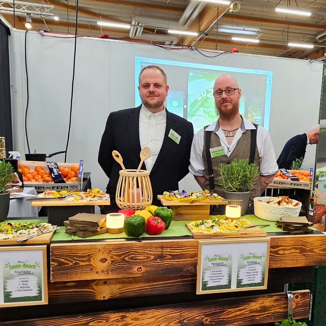 Zwei Mitarbeiter von Manss Foodservice am Messestand mit dekorativen Taste-Boards, Kräutern und frischem Gemüse auf Holztresen