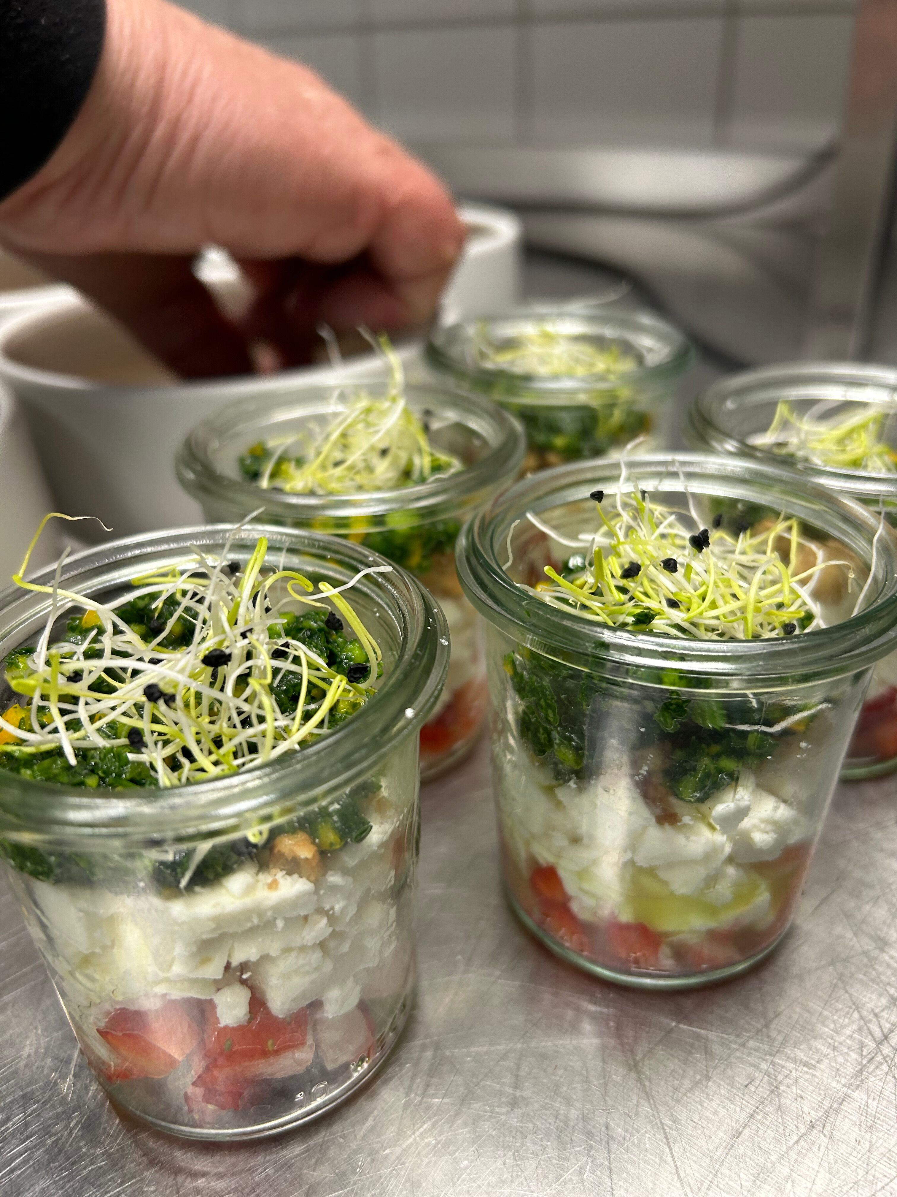 Schichtsalat im Glas mit Feta, Gemüse und Sprossen, Hand im Hintergrund greift nach Geschirr