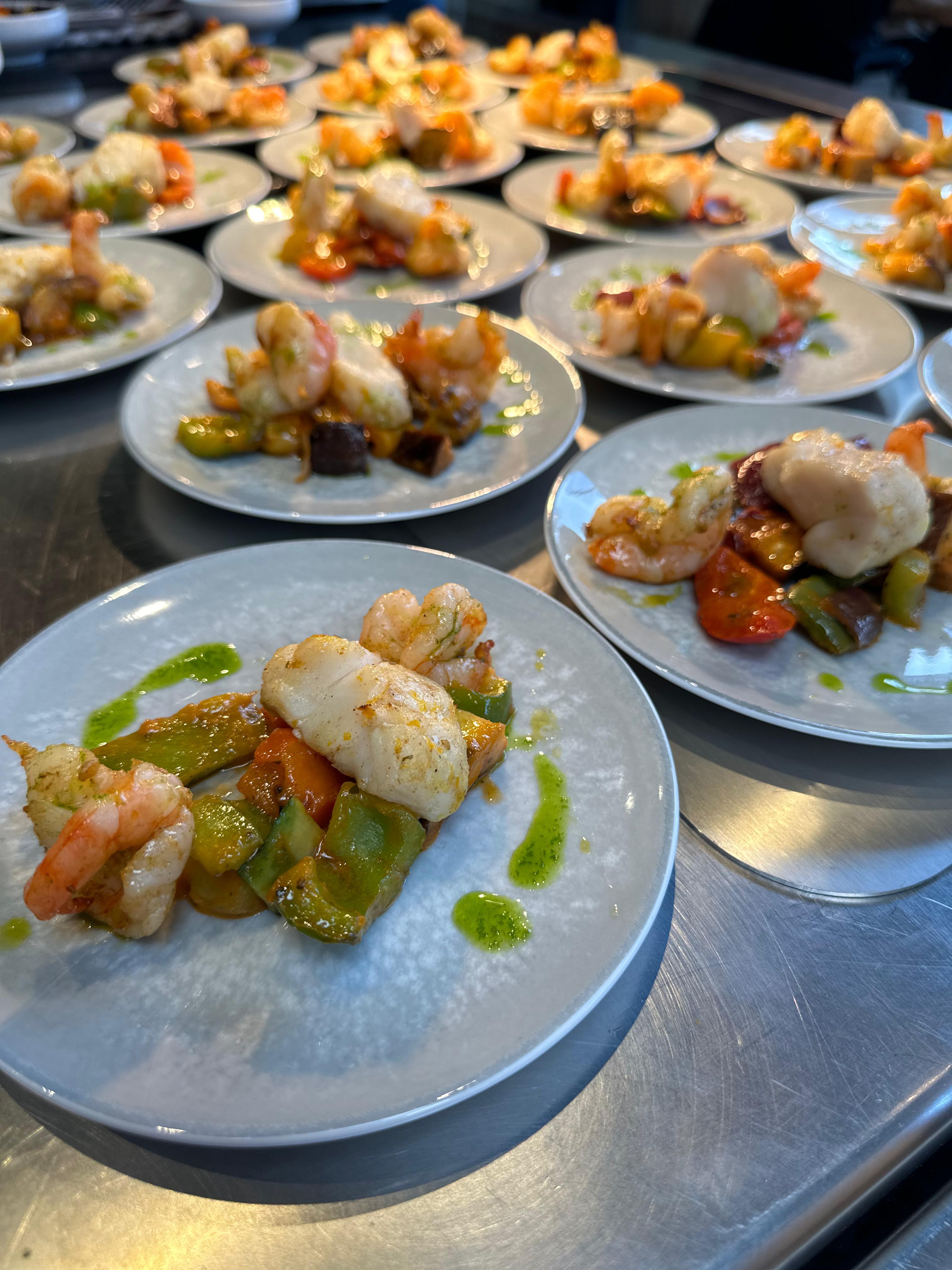 Mehrere angerichtete Teller mit Fischfilet, Garnelen und buntem Gemüse auf Edelstahlfläche – Hauptgang des Frühlingsküche-Workshops von Manss Foodservice