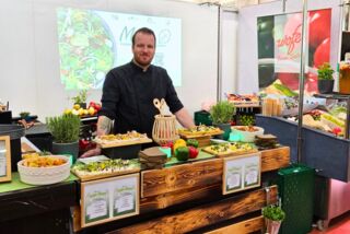 „Ein Koch präsentiert am Stand von Manss Foodservice auf der Ebäko Messe eine Auswahl kreativer Taste-Boards mit frischem Gemüse und Kräutern