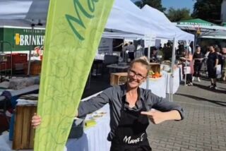 Mitarbeiterin von Manss Foodservice steht lachend auf dem Sommerfest-Gelände neben einer grünen Manss-Fahne, im Hintergrund weiße Pavillons mit Obst- und Gemüseständen