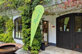 Der Eingangsbereich des Veranstaltungsorts im Sauerland, reich bewachsen mit grünem Efeu und herbstlichen Farbtönen. Vor der offenen Tür steht ein grüner Manss-Foodservice-Aufsteller