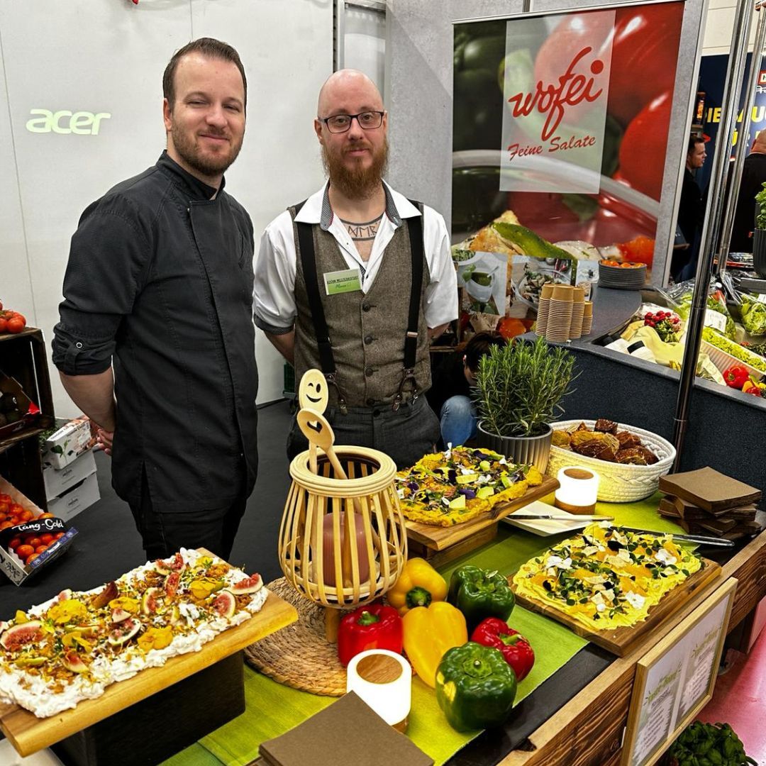 Zwei Teammitglieder von Manss Foodservice stehen am Messestand auf der Ebäko Messe vor bunt belegten Taste-Boards und frischem Gemüse