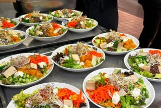 Verschiedene frische Bowls mit bunten Zutaten wie Tofu, Karotten, Sprossen und grünem Salat auf einer Theke, im Hintergrund Mitarbeitende in schwarzer Kleidung.