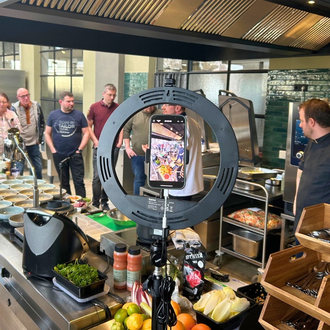Blick in die Showküche beim Manss Workshop am 20. März 2024 – im Vordergrund ein Ringlicht mit Smartphone, im Hintergrund Workshop-Gäste bei einer Präsentation moderner Frühlingsgerichte