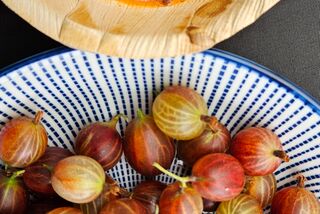 Nahaufnahme von frischen roten Stachelbeeren in einer blauen Schale neben einem Bambusteller mit fruchtig-würzigem Chutney aus Tomaten und Stachelbeeren