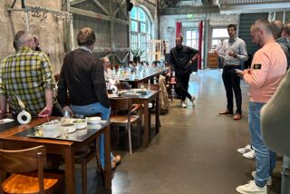 Teilnehmende stehen und sitzen in rustikaler Halle, Einführung durch Workshop-Leitung