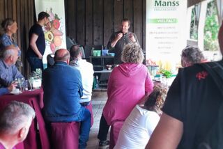 Ein Redner hält im Rahmen einer Waldpilz-Exkursion auf einer überdachten Terrasse einen Vortrag, während die Teilnehmenden aufmerksam zuhören. Im Hintergrund sind Manss-Foodservice-Banner sichtbar.