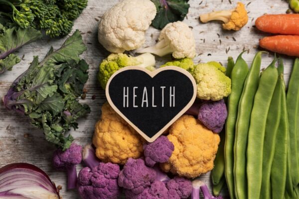 Bunte Auswahl an frischem Gemüse wie Blumenkohl, Karotten, Zwiebeln und Bohnen, dekorativ angerichtet rund um ein Herzschild mit dem Schriftzug ‚Health‘ – Symbolbild für die Gesundheitswoche von Manss Foodservice.