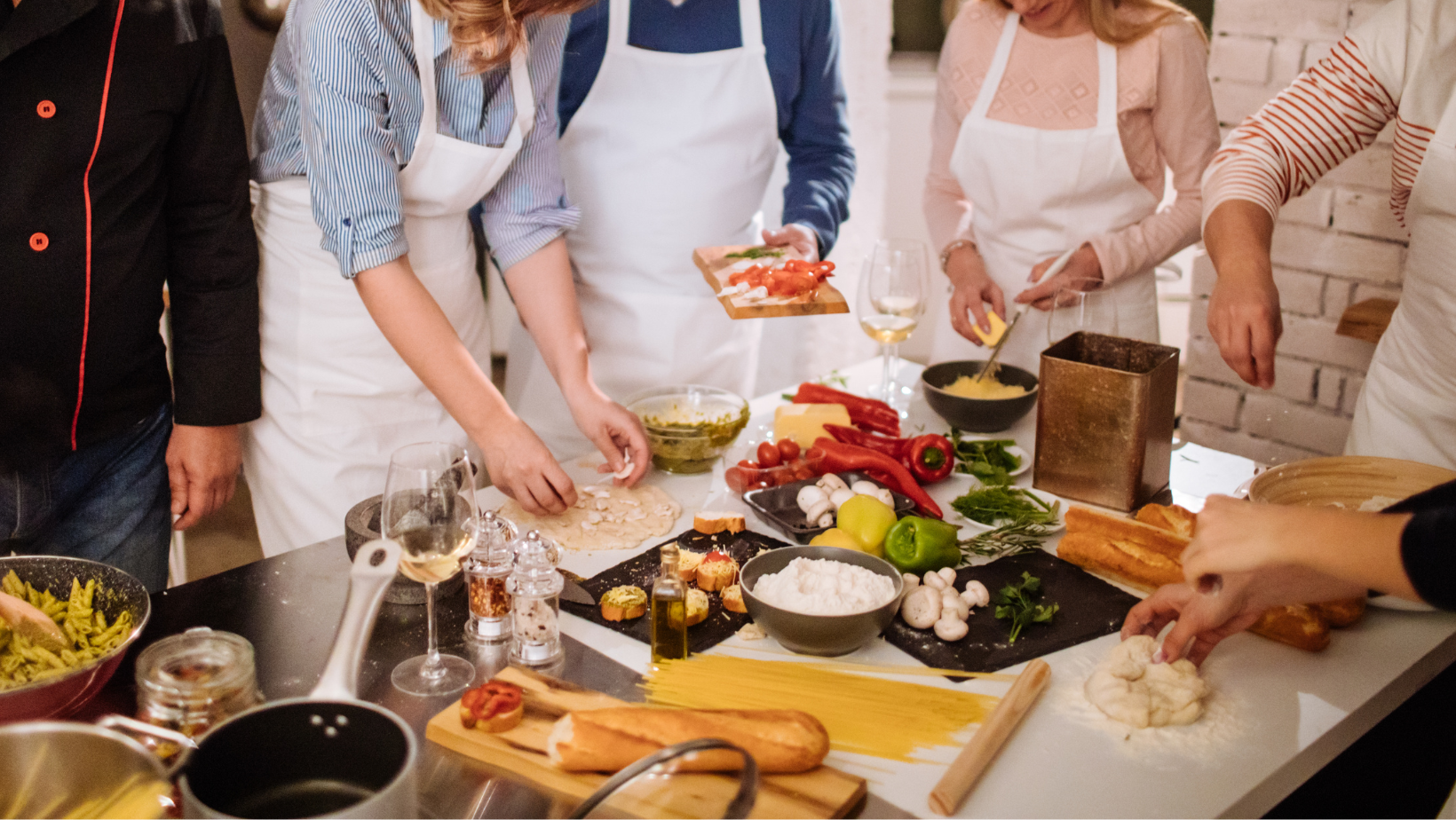 Mehrere Personen mit Schürzen bereiten gemeinsam Speisen in einer modernen Küche zu. Auf dem Tisch liegen frische Zutaten wie Gemüse, Kräuter, Pasta und Baguette. Die Szene vermittelt Geselligkeit und Kochfreude.