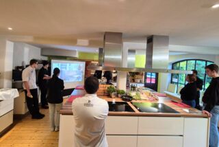 Teilnehmende des Genusslust-Workshops von Manss Foodservice verfolgen eine Kochpräsentation mit Live-Projektion in einer modernen, lichtdurchfluteten Showküche im Sauerland.