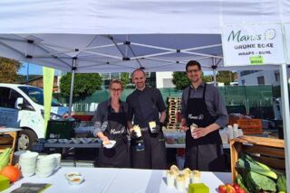 Drei Mitarbeitende von Manss Foodservice präsentieren Wraps und Bowls an einem Stand mit dem Schild ‚Grüne Ecke‘ beim Sommerfest von Blumen Risse und Overkamp