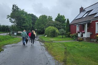 Drei Personen gehen auf einem nassen Weg zwischen Obsthof-Gebäude mit Solardach und grüner Landschaft in Richtung Anbauflächen